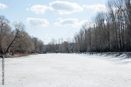Winter panorama of a frozen lake, trees and poles with lanterns in the park.