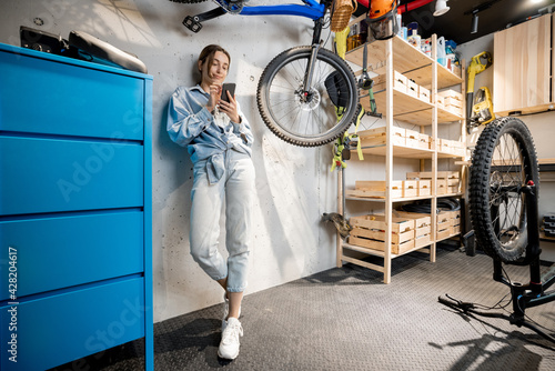 Full length portrait of a young handywoman standing relaxed with phone in the well equipped workshop or garage. DIY concept photo