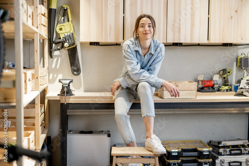 Portrait of a young cheerful handywoman sitting on the workbench in the well equipped workshop at home. DIY conceept photo