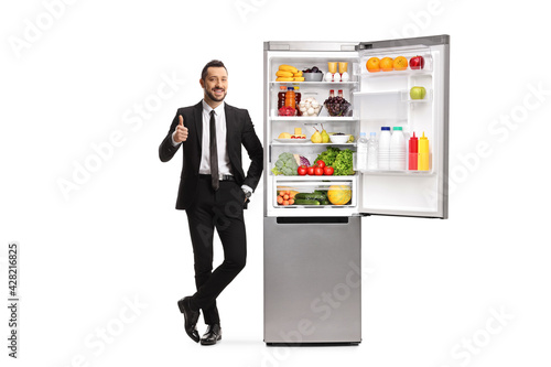 Full length portrait of a young man in a suit leaning on an empty fridge with thumbs up