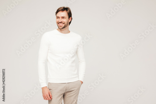 Portrait of handsome smiling hipster lumbersexual businessman model wearing casual white sweater and trousers. Fashion stylish man posing against grey background in studio