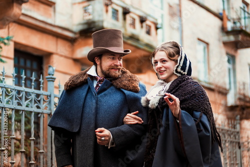 Couple wearing traditional victorian clothes, man and woman walking and talking. Fashion and trends of previous epochs and times. Male wearing cloak, female in dress and hat. 