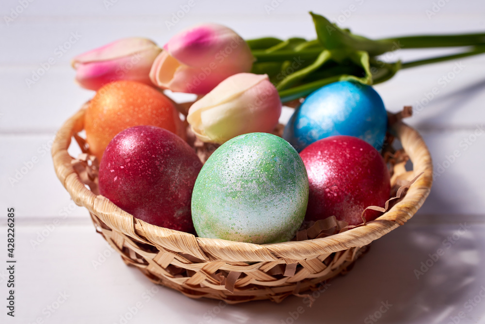 A small basket with colorful painted eggs and a bouquet of tulips on a white wooden background. Orthodox Easter composition. A small basket with colorful painted eggs and a bouquet of tulips on a white wooden background. Orthodox Easter composition.