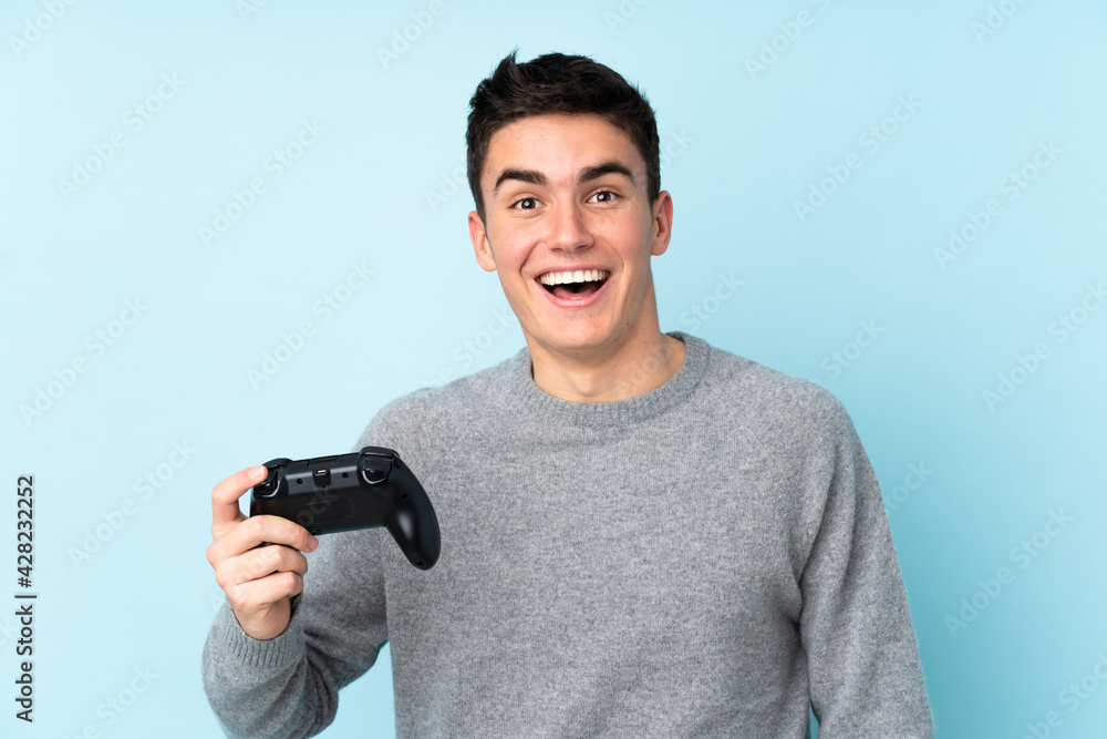 Teenager caucasian man playing with a video game controller isolated on blue background with surprise and shocked facial expression Teenager caucasian man playing with a video game controller isolated on blue background with surprise and shocked facial expression