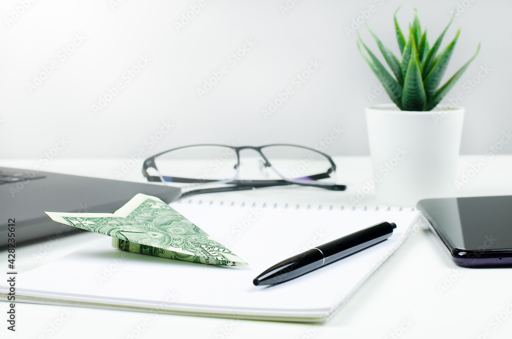 One dollar bill plane on the desk with blank notepad, pen, laptop, smartphone, glasses and green plant. Business concept One dollar bill plane on the desk with blank notepad, pen, laptop, smartphone, glasses and green plant. Business concept