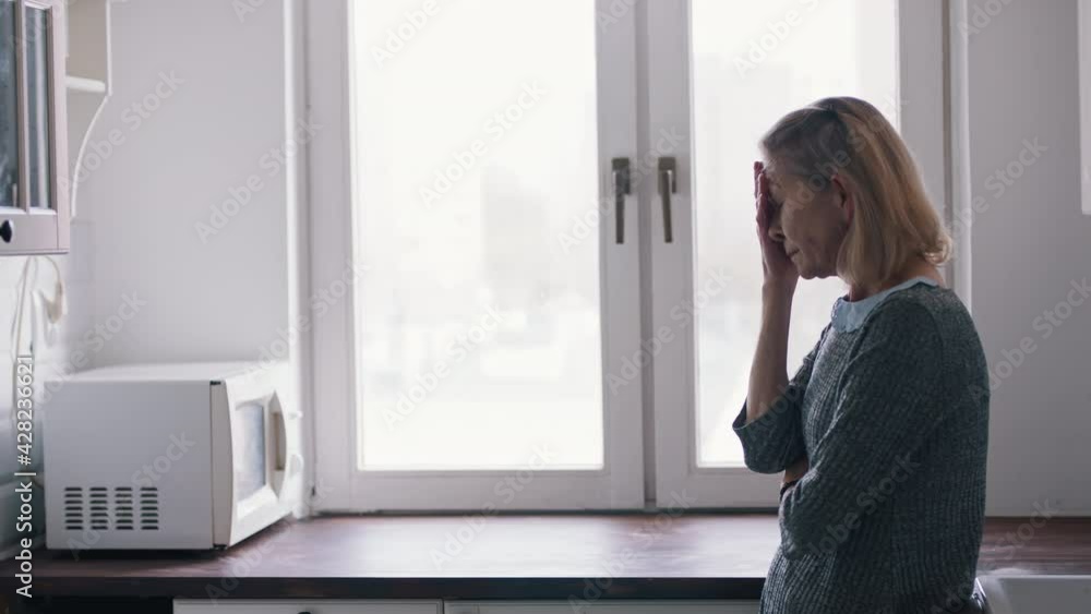 Upset elderly woman holding hand over the forehead while standing in the kitcen. High quality 4k footage Upset elderly woman holding hand over the forehead while standing in the kitcen. High quality 4k footage