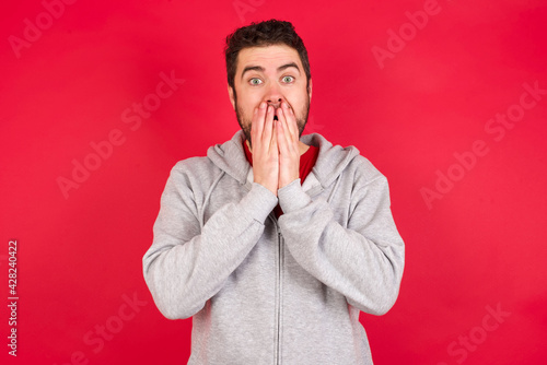 Vivacious Young caucasian man wearing tracksuit over red background , giggles joyfully, covers mouth, has natural laughter, hears positive story or funny anecdote