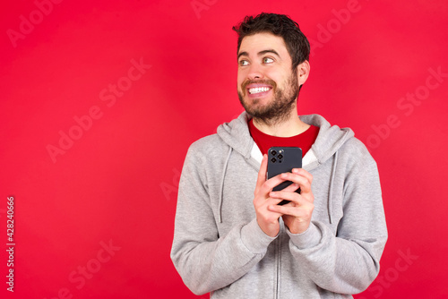 Young caucasian man wearing tracksuit over red background holding a smartphone and looking sideways at blank copyspace. © Roquillo