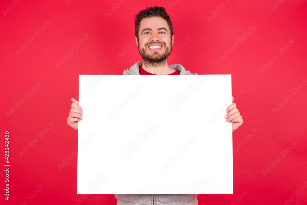 Happy Young caucasian man wearing tracksuit over red background Holding Empty Paper Board Advertising Offer Text Standing. Autumn Advertisement Banner. Happy Young caucasian man wearing tracksuit over red background Holding Empty Paper Board Advertising Offer Text Standing. Autumn Advertisement Banner.