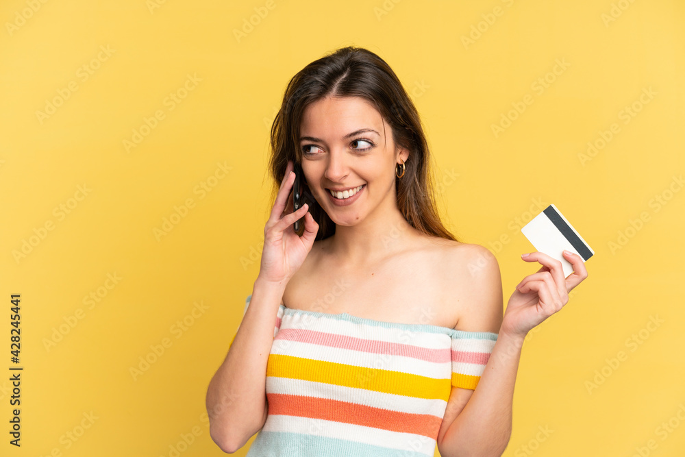 Young caucasian woman isolated on yellow background keeping a conversation with the mobile phone and holding a credit card Young caucasian woman isolated on yellow background keeping a conversation with the mobile phone and holding a credit card