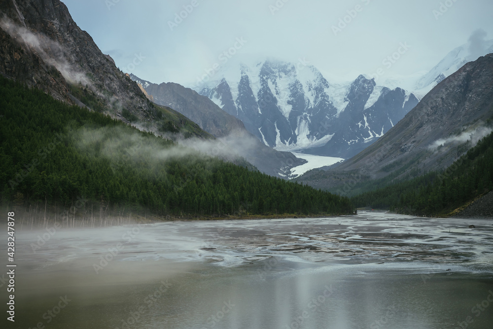 Atmospheric alpine landscape with mountain lake with streams from snowy mountains in overcast weather. Gloomy mountain scenery with green lake with rainy circles and low clouds in mountain valley.