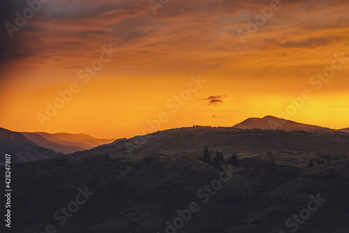 Atmospheric landscape with silhouettes of mountains and hills with trees on background of orange dawn sky. Colorful nature scenery with sunset or sunrise of illuminating color. Sundown paysage.