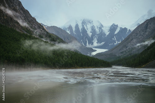 Atmospheric alpine landscape with mountain lake with streams from snowy mountains in overcast weather. Gloomy mountain scenery with green lake with rainy circles and low clouds in mountain valley.