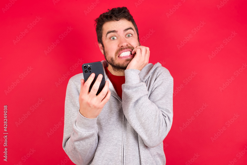 Afraid funny Young caucasian man wearing tracksuit over red background holding telephone and bitting nails Afraid funny Young caucasian man wearing tracksuit over red background holding telephone and bitting nails