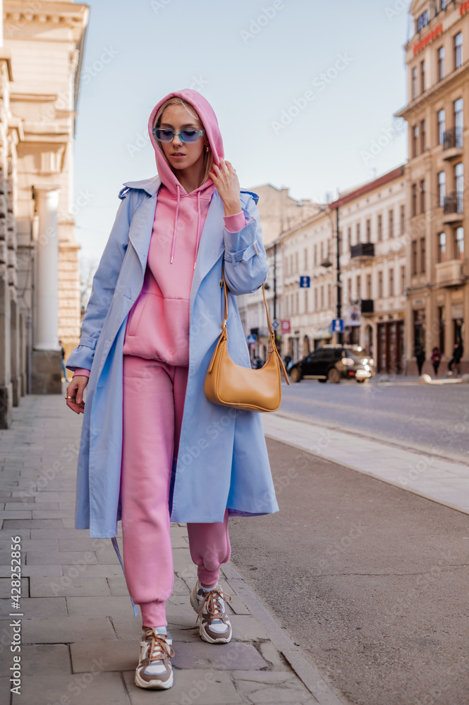 Streetstyle, street fashion concept: woman wearing trendy sport chic style  outfit walking in city. Blue trench coat, sunglasses, pink hoodie,  trousers, sneakers, yellow bag Stock Photo | Adobe Stock