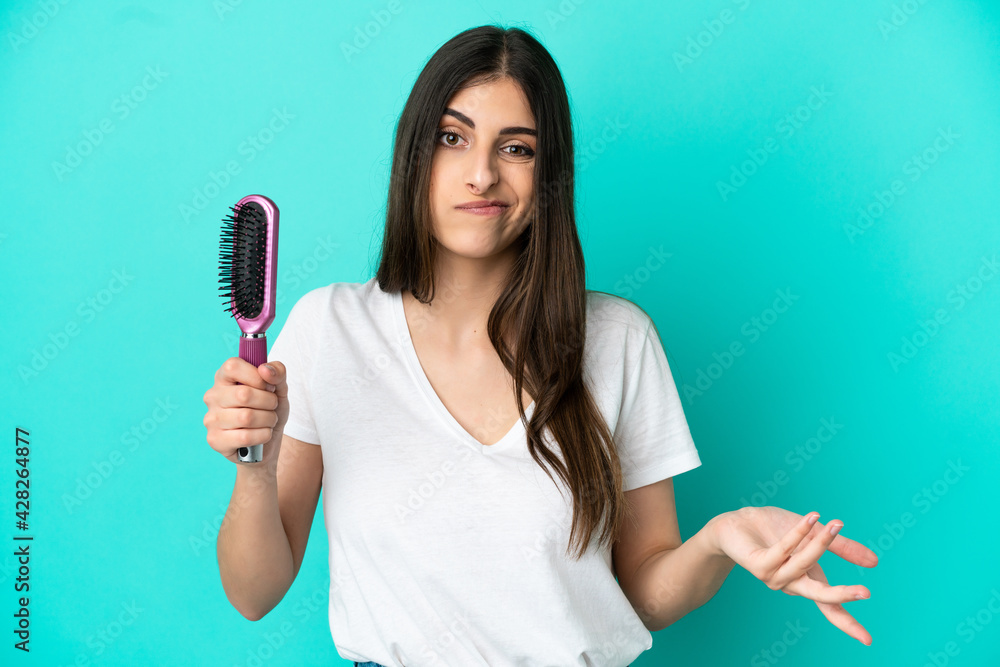 Young caucasian woman with hair comb isolated on blue background making doubts gesture while lifting the shoulders Young caucasian woman with hair comb isolated on blue background making doubts gesture while lifting the shoulders