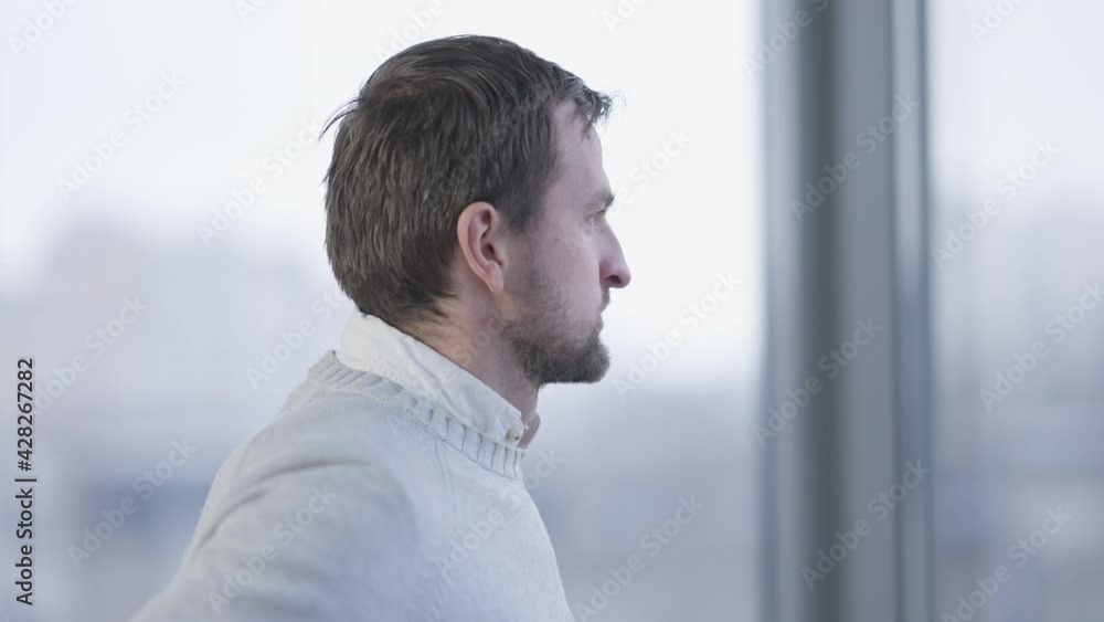Side view of anxious thoughtful Caucasian man looking out the window in office. Portrait of worried young bearded brunette CEO manager thinking on project idea indoors. Management and business Side view of anxious thoughtful Caucasian man looking out the window in office. Portrait of worried young bearded brunette CEO manager thinking on project idea indoors. Management and business