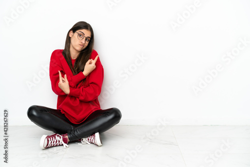 Young woman sitting on the floor isolated on white background pointing to the laterals having doubts