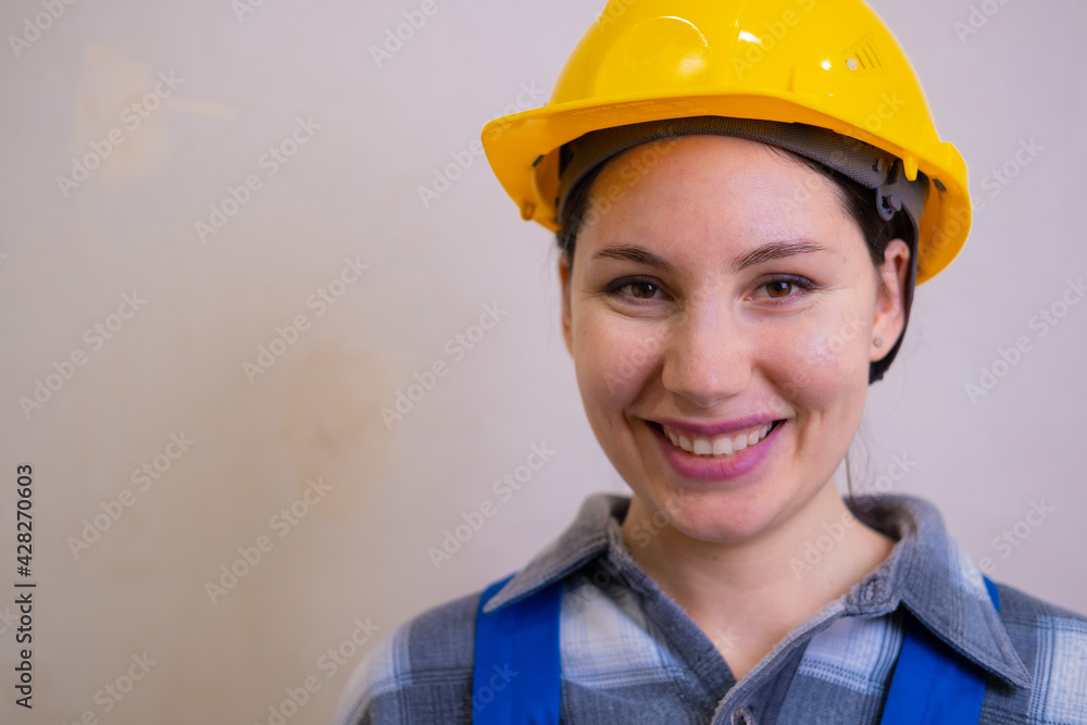 A beautiful construction worker girl in a yellow hard hat and a checkered shirt looks at the camera, smiles and winks against the background of a stucco wall. A beautiful construction worker girl in a yellow hard hat and a checkered shirt looks at the camera, smiles and winks against the background of a stucco wall.