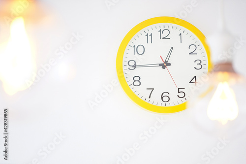 White round clock with yellow frame on a white wall and blurred lamp decorations.