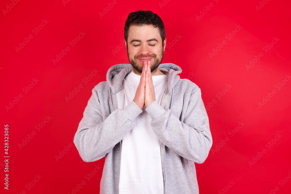 Indoor closeup of young handsome caucasian man in sports clothes against red wall practicing yoga and meditation, holding palms together in namaste, looking calm, relaxed and peaceful. Indoor closeup of young handsome caucasian man in sports clothes against red wall practicing yoga and meditation, holding palms together in namaste, looking calm, relaxed and peaceful.