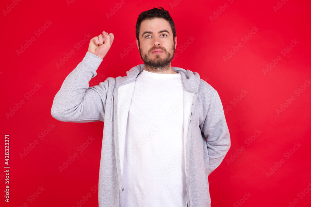 young handsome caucasian man in sports clothes against red wall feeling serious, strong and rebellious, raising fist up, protesting or fighting for revolution. young handsome caucasian man in sports clothes against red wall feeling serious, strong and rebellious, raising fist up, protesting or fighting for revolution.