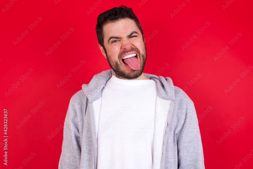 young handsome caucasian man in sports clothes against red wall with happy and funny face smiling and showing tongue. young handsome caucasian man in sports clothes against red wall with happy and funny face smiling and showing tongue.