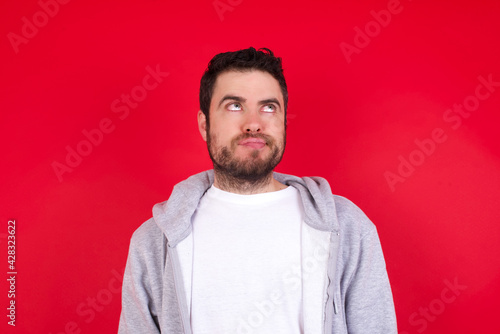 young handsome caucasian man in sports clothes against red wall has worried face looking up lips together, being upset thinking about something important, keeps hands down.
