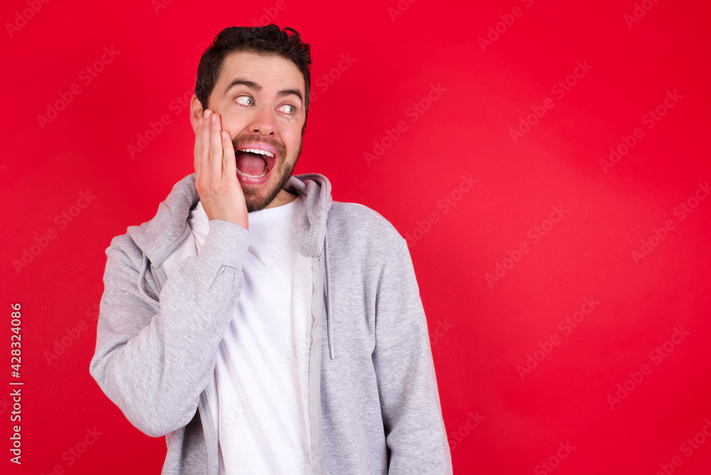 young handsome caucasian man in sports clothes against red wall excited looking to the side hand on face. Advertisement and amazement concept. young handsome caucasian man in sports clothes against red wall excited looking to the side hand on face. Advertisement and amazement concept.
