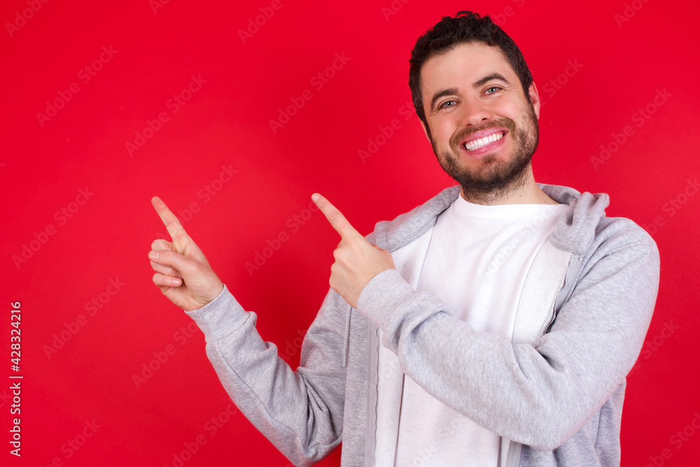 young handsome caucasian man in sports clothes against red wall with positive expression, indicates with fore finger at blank copy space for your promotional text or advertisement. young handsome caucasian man in sports clothes against red wall with positive expression, indicates with fore finger at blank copy space for your promotional text or advertisement.