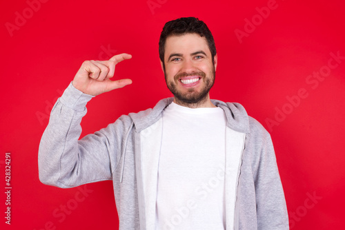young handsome caucasian man in sports clothes against red wall smiling and gesturing with hand small size, measure symbol.