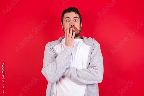 young handsome caucasian man in sports clothes against red wall covering mouth with hands scared from something or someone bitting nails © Jihan