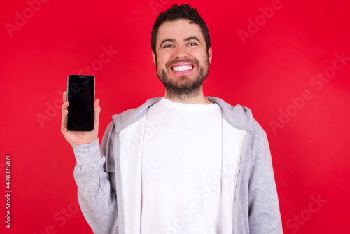 Smiling young handsome caucasian man in sports clothes against red wall showing empty phone screen. Advertisement and communication concept.