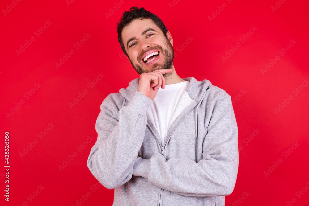 young handsome caucasian man in sports clothes against red wall laughs happily keeps hand on chin expresses positive emotions smiles broadly has carefree expression young handsome caucasian man in sports clothes against red wall laughs happily keeps hand on chin expresses positive emotions smiles broadly has carefree expression