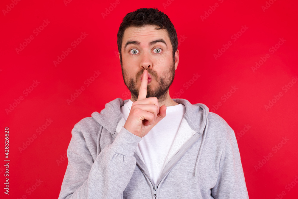 Surprised young handsome caucasian man in sports clothes against red wall makes silence gesture, keeps finger over lips and looks mysterious at camera Surprised young handsome caucasian man in sports clothes against red wall makes silence gesture, keeps finger over lips and looks mysterious at camera