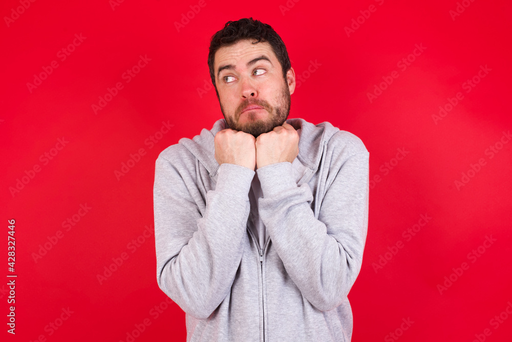 Curious young handsome caucasian man in sports clothes against red background keeps hands under chin bites lips and looks with interest aside. Curious young handsome caucasian man in sports clothes against red background keeps hands under chin bites lips and looks with interest aside.