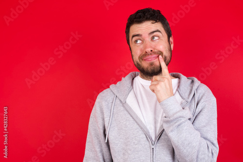 Lovely dreamy young handsome caucasian man in sports clothes against red background keeps finger near lips looks aside copy space.