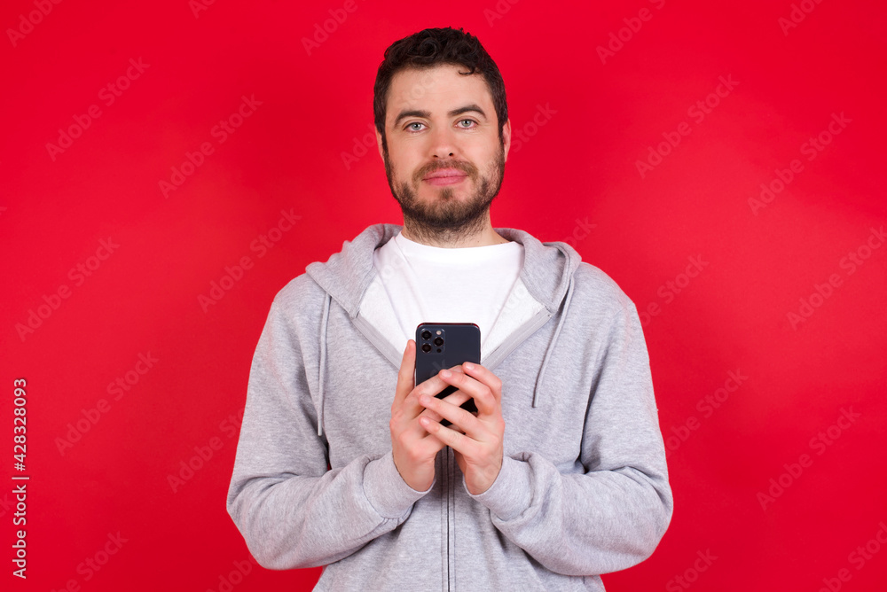 Portrait of serious confident young handsome caucasian man in sports clothes against red background holding phone in two hands Portrait of serious confident young handsome caucasian man in sports clothes against red background holding phone in two hands