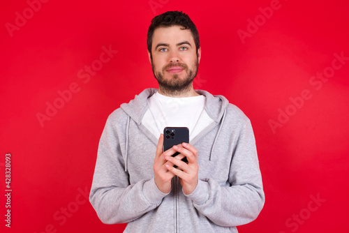 Portrait of serious confident young handsome caucasian man in sports clothes against red background holding phone in two hands © Jihan