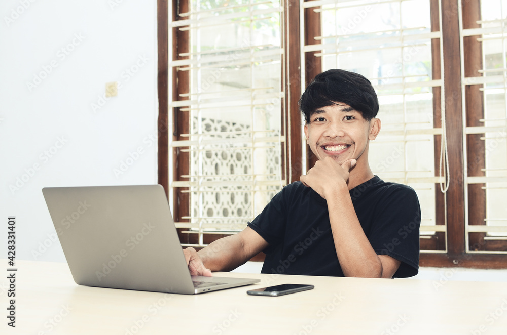 The Asian man who is sitting at the office table wearing a black shirt is smiling in front of the laptop