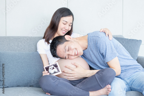 Father is kissing his mother's stomach. Young husband kissing his pregnant wife's tummy in living room with the baby ultrasound photo.
