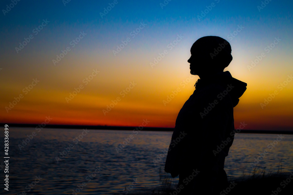Fototapeta premium The girl at sunset stands at the shore and looks into the distance, waiting for a miracle.