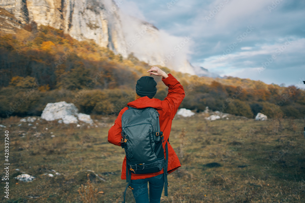 woman in a gray hat red jacket with a backpack gestures with her hands on nature in the mountains