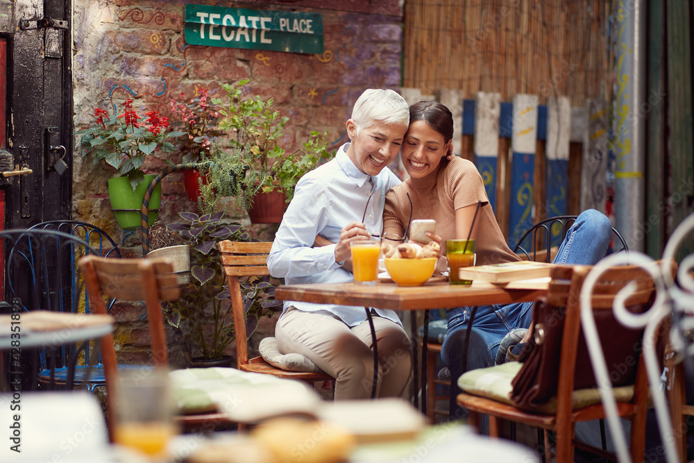 An older woman and her young female friend are enjoying a smartphone content while they have a drink in the bar. Leisure, bar, friendship, outdoor An older woman and her young female friend are enjoying a smartphone content while they have a drink in the bar. Leisure, bar, friendship, outdoor