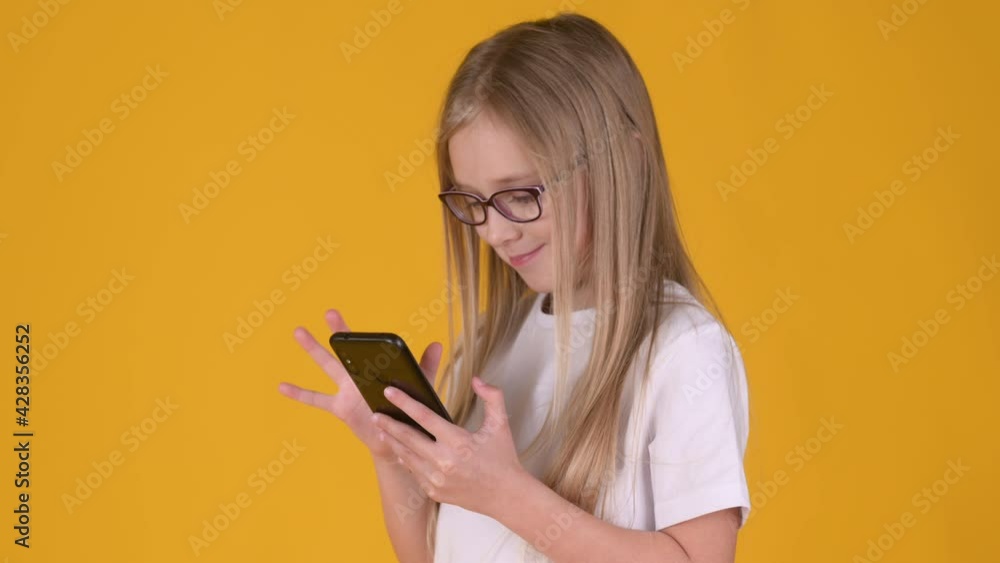 Little blonde girl in white T-shirt and glasses Using mobile cell phone isolated on yellow background. Childhood lifestyle concept. Little blonde girl in white T-shirt and glasses Using mobile cell phone isolated on yellow background. Childhood lifestyle concept.