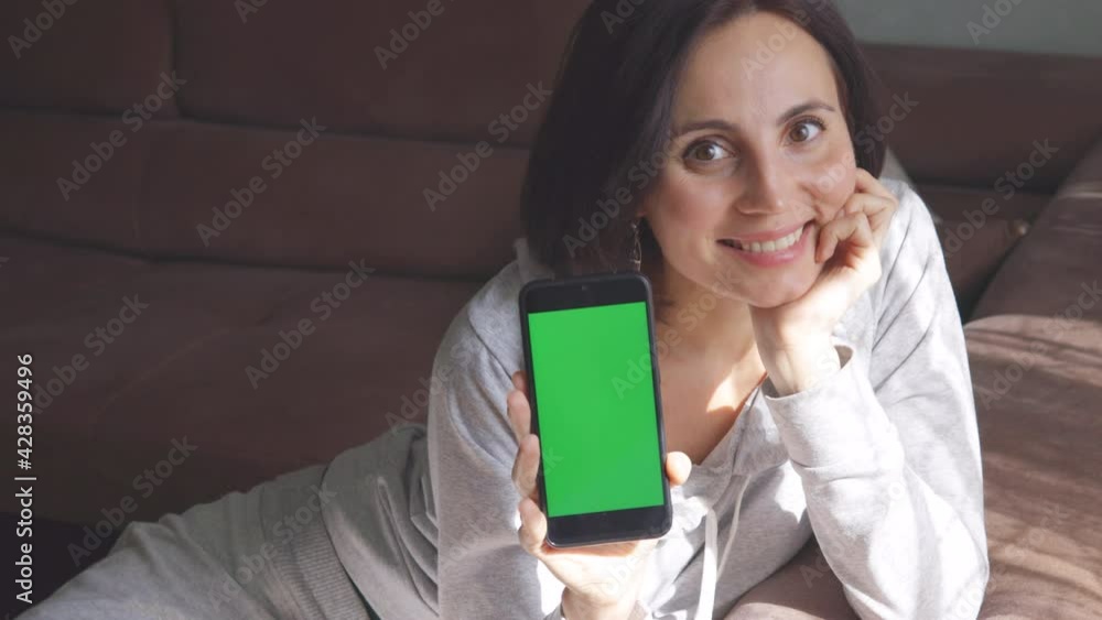 European woman with short hair in grey clothes sitting on comfy sofa at home smiling and showing a mobile phone with green screen. Good for branding application European woman with short hair in grey clothes sitting on comfy sofa at home smiling and showing a mobile phone with green screen. Good for branding application