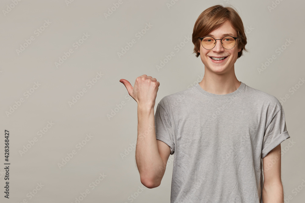 Portrait of handsome, adult male with blond hair. Wearing grey t-shirt, glasses and has braces. Watching at the camera and pointing with thumb to the left at copy space, isolated over grey background Portrait of handsome, adult male with blond hair. Wearing grey t-shirt, glasses and has braces. Watching at the camera and pointing with thumb to the left at copy space, isolated over grey background