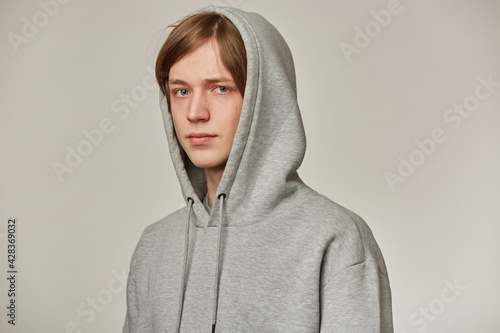 Cool looking male, handsome guy with blond hair. Wearing grey hoodie and puts hood on. People and emotion concept. Watching serious at the camera isolated over grey background © timtimphoto