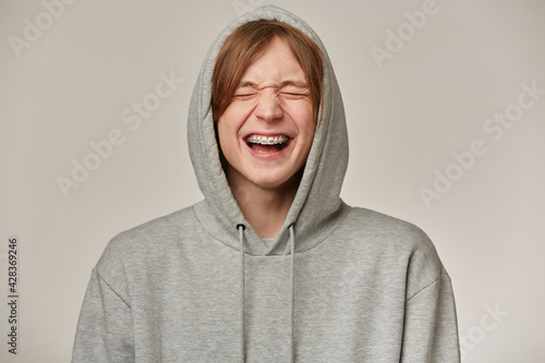 Cheerful male, handsome guy with blond hair. Wearing grey hoodie. Has braces. People and emotion concept. Puts hood on and laughing with closed eyes. Stand isolated over grey background © timtimphoto