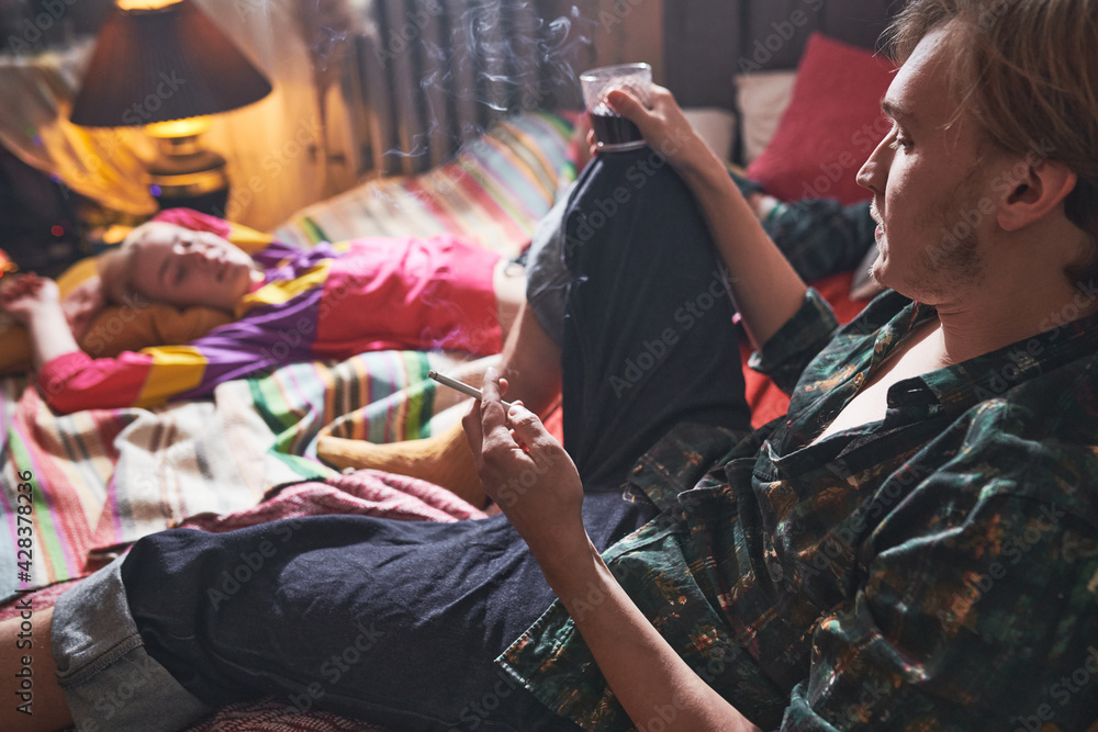 Fototapeta premium Young man smoking a cigarette and drinking wine sitting on the bed with his girlfriend relaxing near by him at home